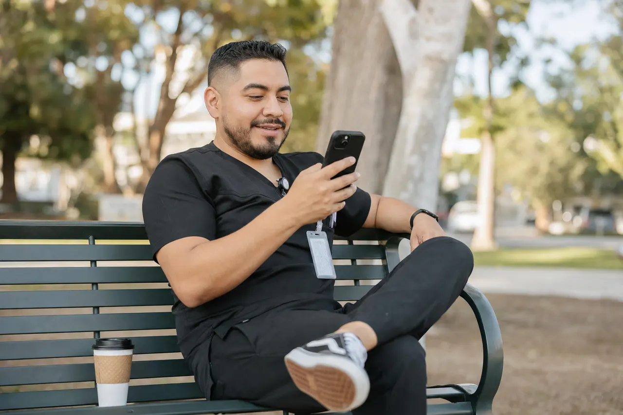 A support worker in scrubs sits on a park bench, checking his phone.
