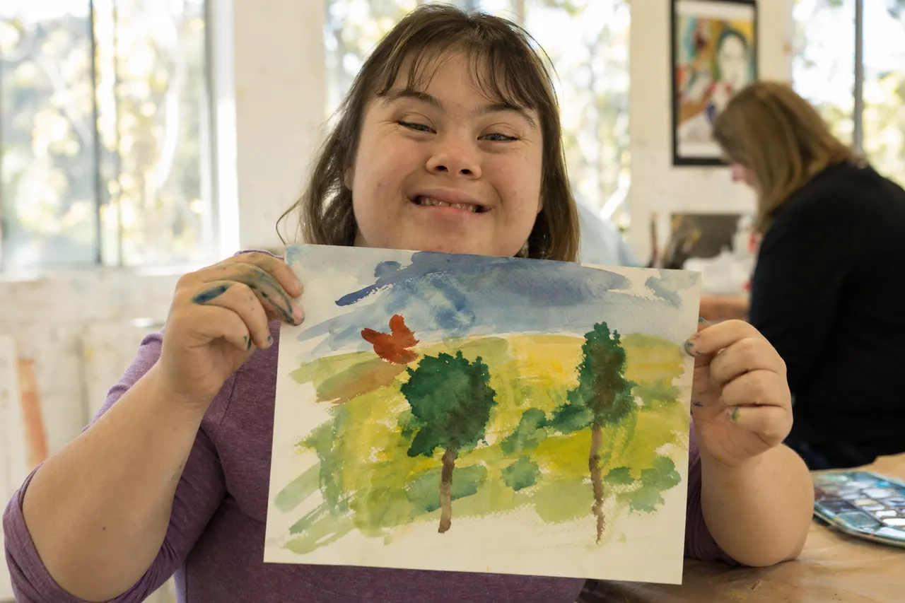 A smiling young woman holds up her watercolor painting in an art class.